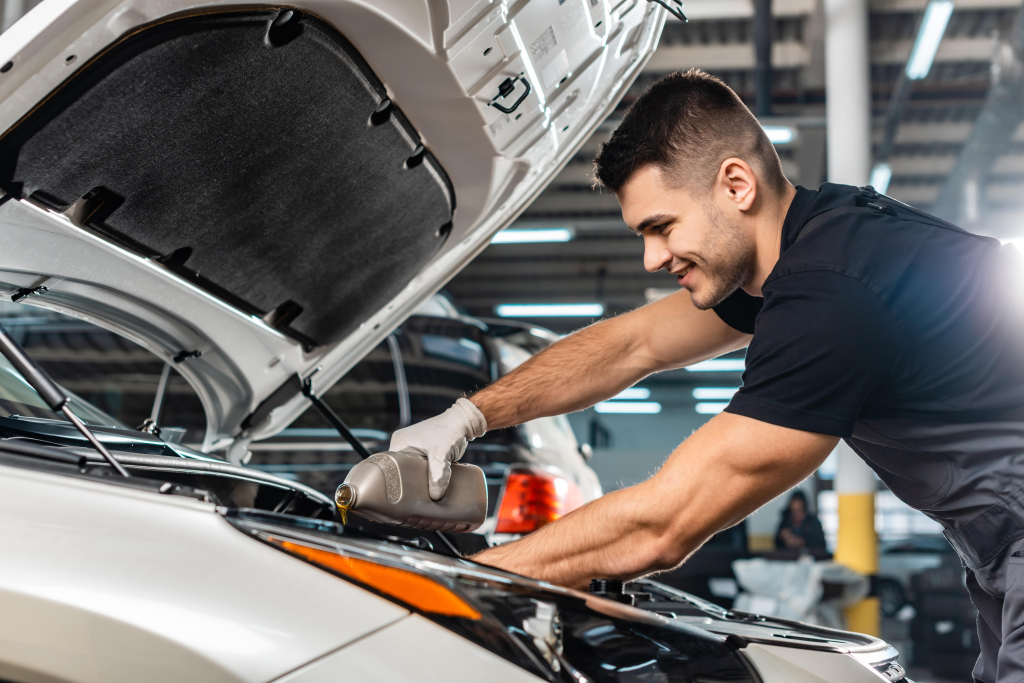 A mechanic with a smile pours oil into a car engine in a brightly lit garage. The hood is open, and other cars are visible in the background.