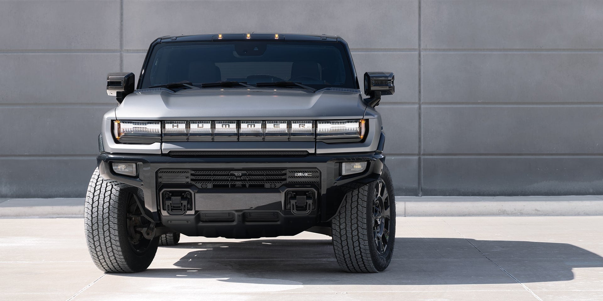 Front view of a matte grey GMC Hummer EV truck parked against a concrete wall.