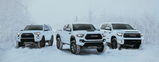 Three white Toyota trucks are parked on a snowy road surrounded by frost-covered trees. The scene conveys power and durability in a winter setting.