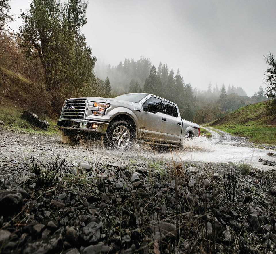 A silver Ford pickup truck drives through a muddy, rocky trail in a forested area under a gray sky, splashing water dynamically, conveying adventure.