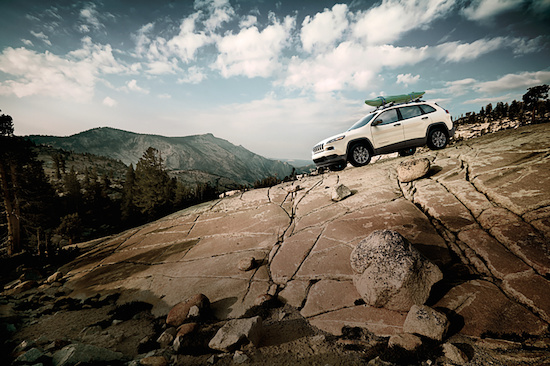 A white SUV with surfboards on top is parked on a rocky, sloped terrain under a cloudy sky. Mountainous landscape and coniferous trees in the background.