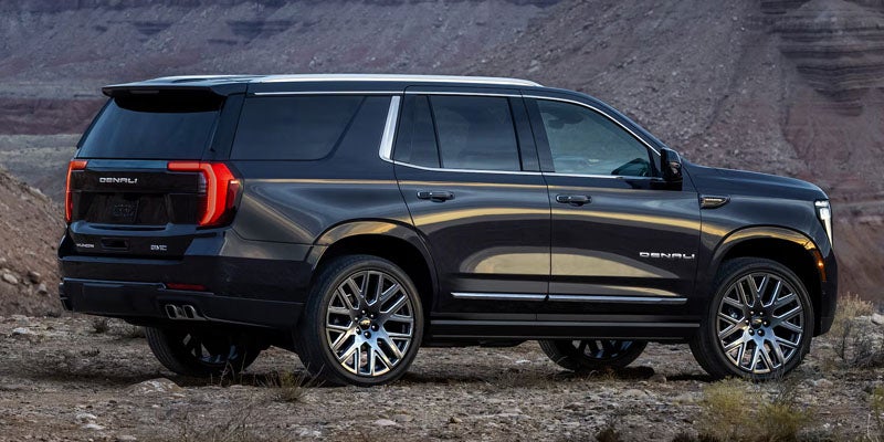 Rear-three-quarter view of a dark gray GMC Yukon Denali parked on rocky, dirt terrain with desert mountains in the background.