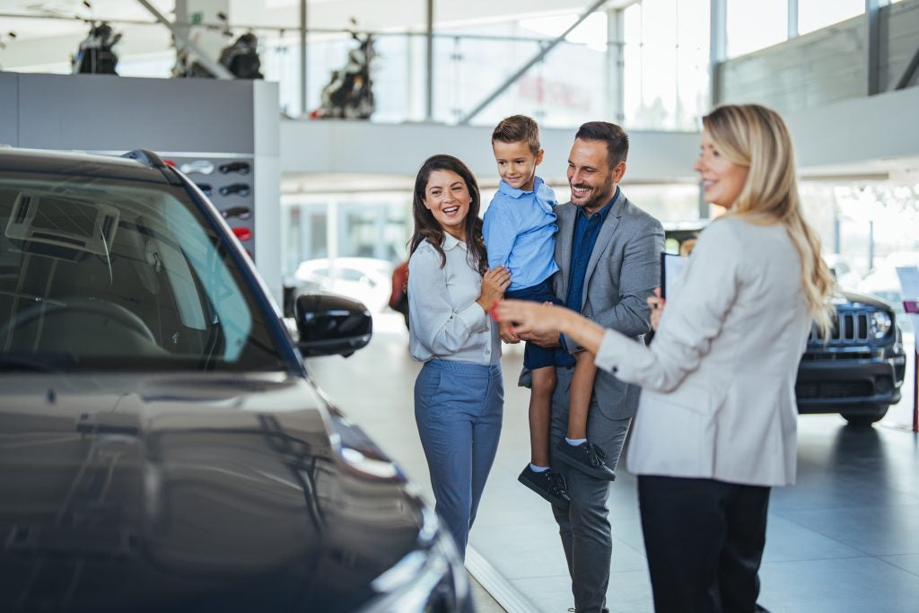 A smiling family of three looking at a new vehicle in a spacious car showroom while a female sales representative points toward the car.