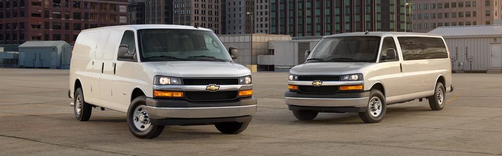 Two white Chevrolet cargo vans parked in an urban lot with city buildings in the background.