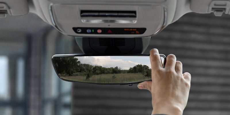A hand adjusting the rearview mirror in a 2025 GMC Terrain Elevation, showing trees and field in the reflection.