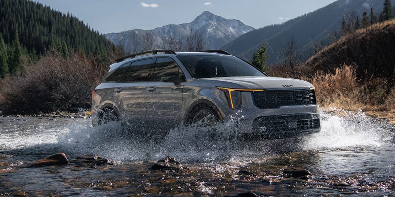 A silver Kia Sorento drives through a shallow mountain stream, splashing water. Surrounding scenery includes lush trees, bushes, and a distant snowy peak.