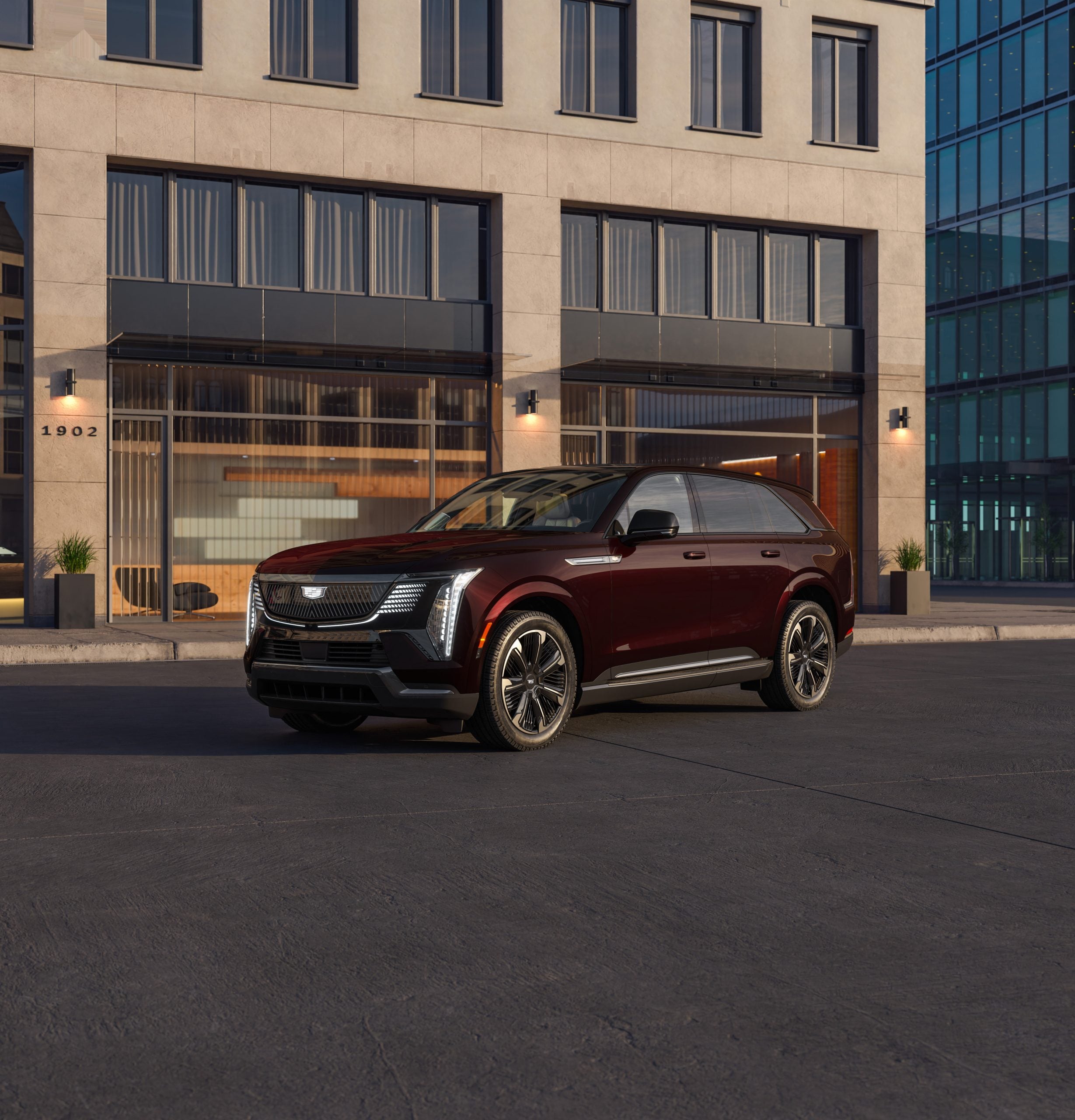 A luxurious maroon 2025 Cadillac ESCALADE IQ SUV parked in front of a modern building with large windows.