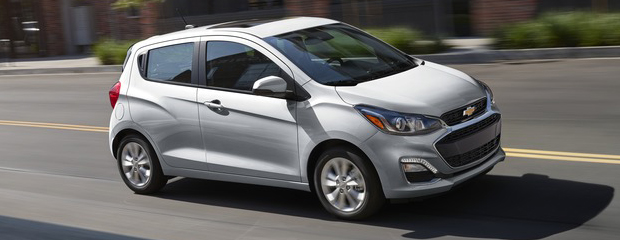 A silver compact car drives on a sunny urban street. The background includes blurred buildings and greenery, conveying speed and modernity.