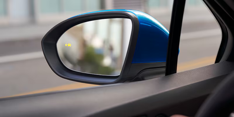 View through a car's blue side mirror showing a blurred city street. A yellow warning icon is visible, indicating a vehicle in the blind spot.