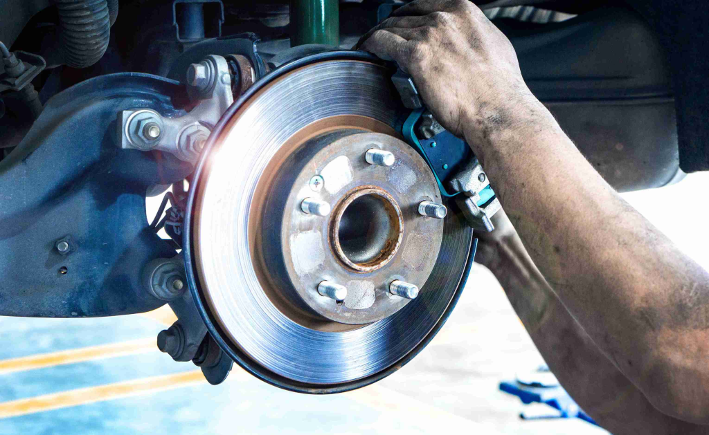 Mechanic working on a car's brake system, focusing on the shiny rotor. Hands are dirty, conveying hands-on labor. Bright, detailed imagery.