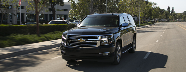 A black SUV drives on a sunny, tree-lined suburban street with modern buildings in the background. The scene conveys a sense of power and modernity.