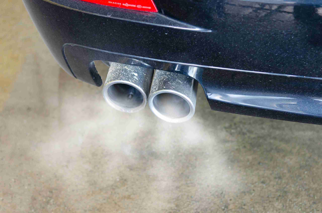 Close-up of a car's dual exhaust pipes emitting white smoke, suggesting the vehicle is running. The background is a concrete surface.