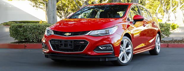 A shiny red Chevrolet Cruze hatchback is parked on a sunny day, surrounded by green shrubs and trees, showcasing a sleek, modern design.