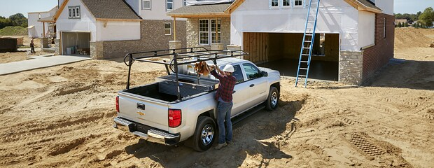 A construction worker, standing near a silver pickup truck, unloads tools at a sandy building site with partially built houses and a blue ladder.