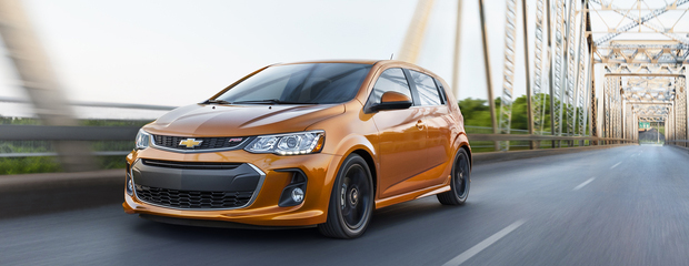 An orange hatchback car drives on a modern steel bridge, showcasing sleek design and speed. The background features greenery and a blue sky.
