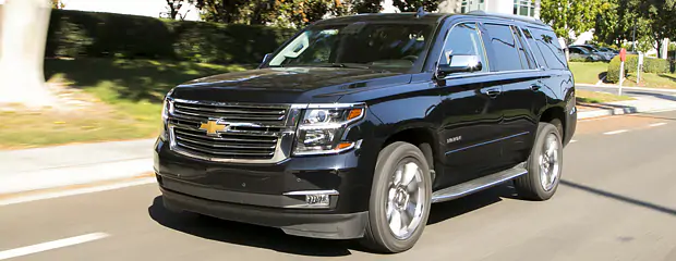 A black Chevrolet SUV drives along a sunny suburban street, with trees and grass visible in the background. The vehicle conveys a sense of power and elegance.