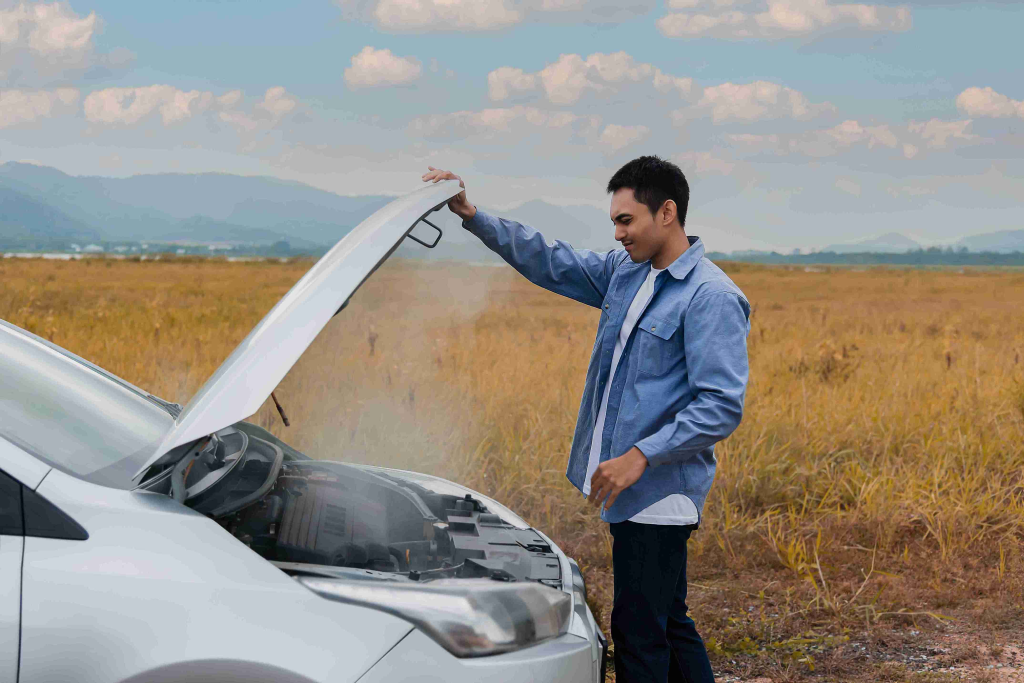 A man in a blue shirt inspects a white car's smoking engine, set against a backdrop of a grassy field and cloudy sky, conveying concern and focus.