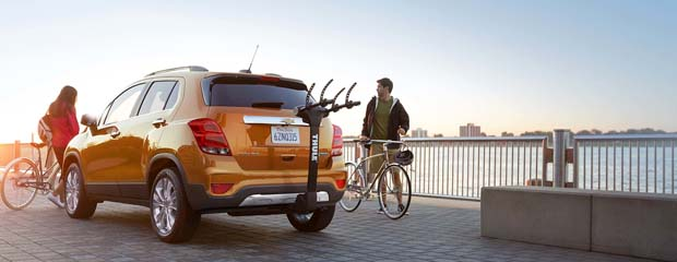 A yellow SUV with a rear bike rack is parked near a waterfront. A man and a woman stand with bicycles, enjoying the scenic view under a clear sky.