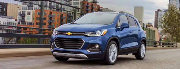 Blue Chevrolet Trax SUV driving on a city road, with modern buildings in the background. The scene conveys urban mobility and modernity.