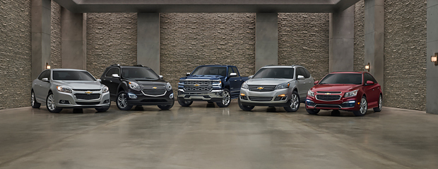 Five Chevrolet vehicles are parked in a modern showroom with concrete walls. The cars are silver, black, blue, gray, and red, lined up side by side.