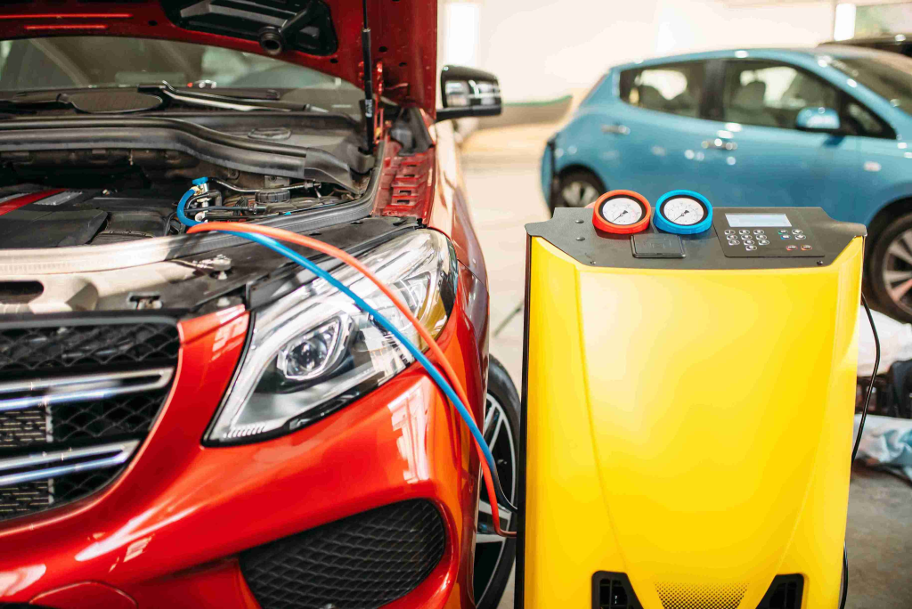 Red car with open hood being serviced, connected by blue and red hoses to a yellow machine with pressure gauges, inside a garage.