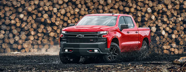 Red pickup truck driving through muddy terrain with stacked logs in the background. The scene conveys ruggedness and strength, suggesting off-road capability.