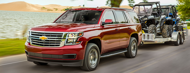 Red SUV towing two off-road vehicles on a trailer, driving by a lake with sand dunes and trees. The scene conveys a sense of adventure and capability.