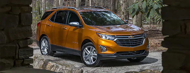 Orange SUV parked on a gravel path in a wooded area. The car has a sleek design with shiny chrome accents, conveying a sense of adventure and style.