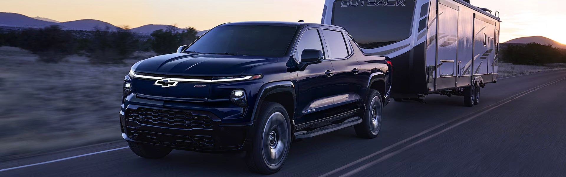 Black 2025 Chevrolet Silverado EV towing a large Outback travel trailer on an open highway at sunset.