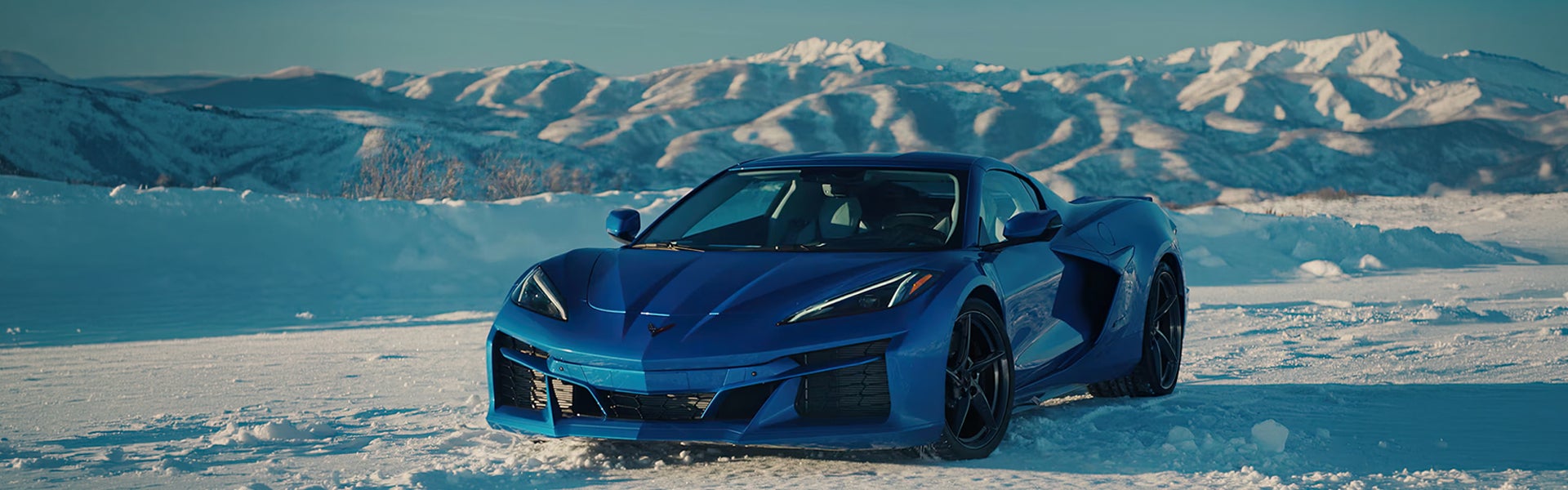 A blue 2026 Chevrolet Corvette E-Ray parked in snow with mountains in the background.