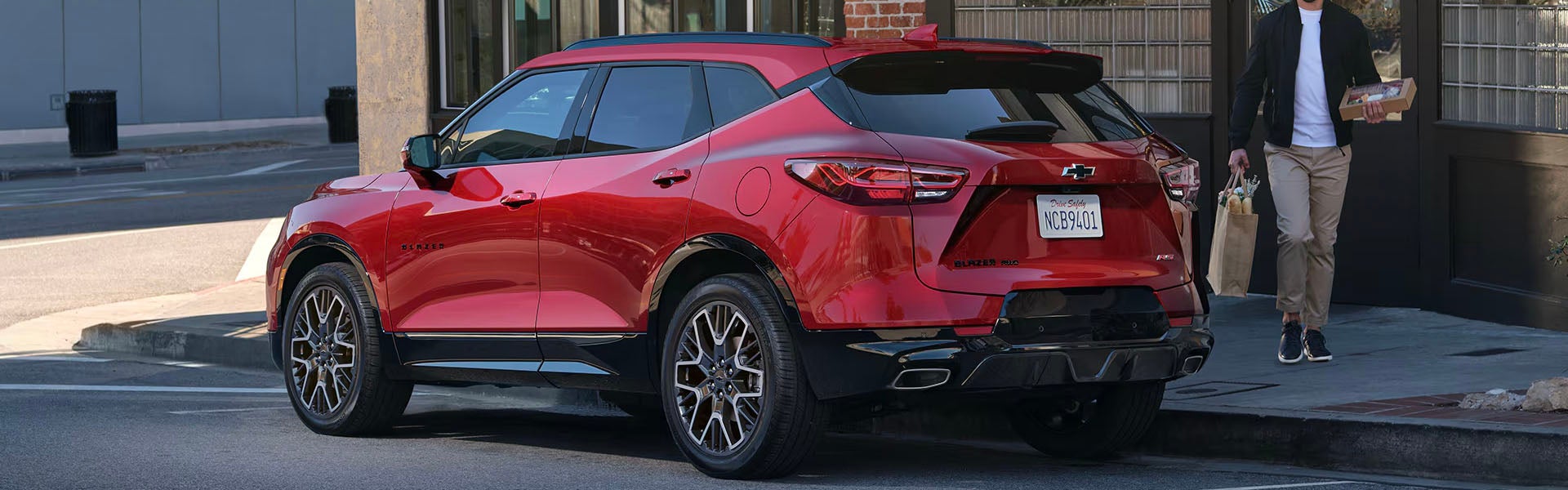 2025 Chevrolet Blazer in red shown from a rear three-quarter angle, featuring sporty LED taillights, black roof accents, alloy wheels, and modern SUV styling parked on a city street near storefronts.