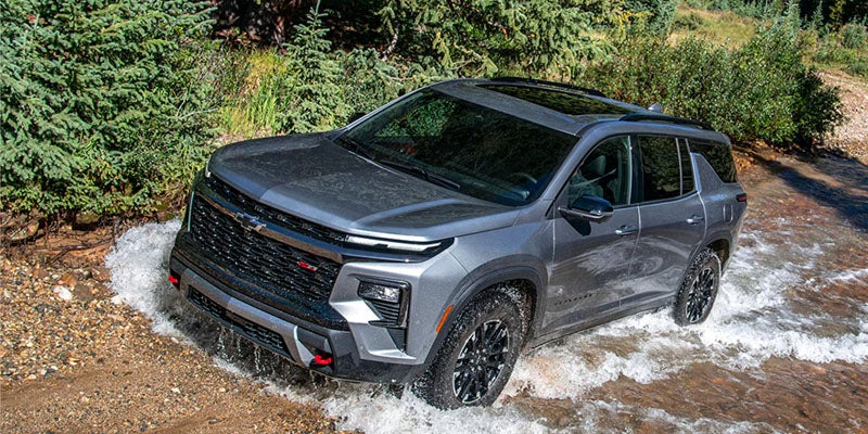 Gray 2026 Chevrolet SUV driving through a shallow stream, shown off-road in a forested area.