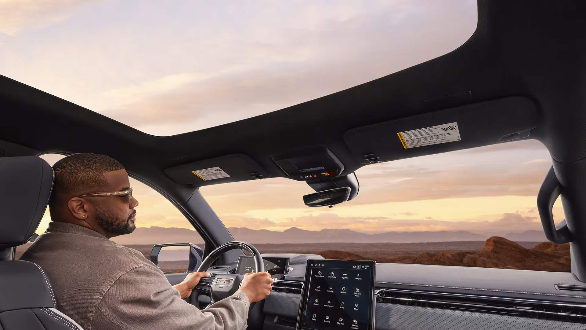 Driver inside GMC Sierra EV with panoramic glass roof and large touchscreen display overlooking desert landscape at sunset.