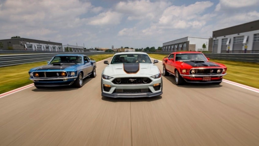 New and vintage Ford Mustangs in a row driving on a race track