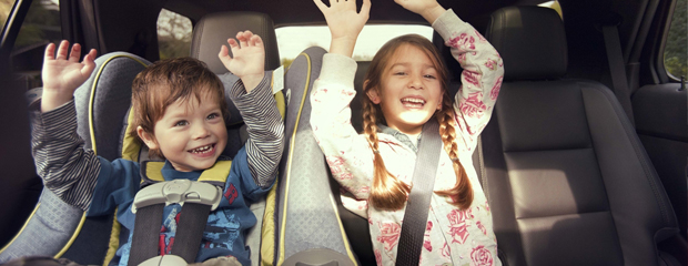 Two children smiling in a car, one in a car seat and the other with hands raised. They appear joyful and excited, enjoying the ride together.