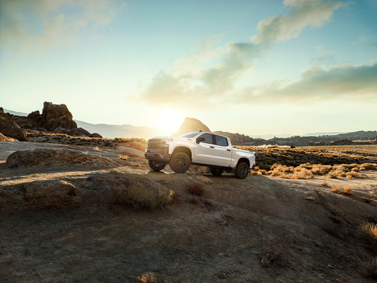 A white pickup truck is parked on rocky terrain at sunrise, with rugged mountains and a partly cloudy sky in the background, conveying adventure.