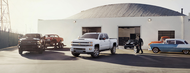 Several Chevrolet trucks and classic cars are parked outside a white industrial garage with a rounded roof. The setting has a sunny, nostalgic tone.