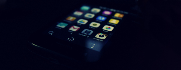 Close-up of a smartphone screen in a dark setting, displaying brightly lit app icons like Instagram and Gmail, creating a moody, focused atmosphere.