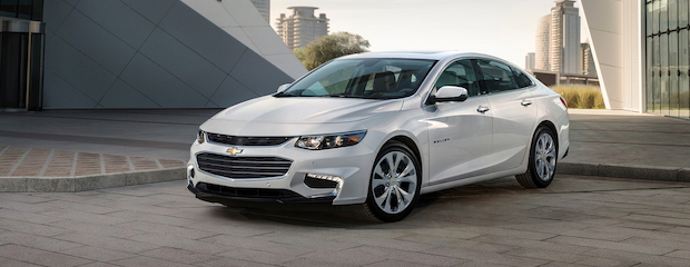 A sleek, white sedan is parked on a modern city street with geometric architecture and skyscrapers in the background, conveying sophistication and style.