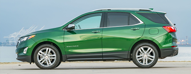 A shiny green SUV is parked on a concrete surface near a waterfront. The background features cranes against a pale blue sky, conveying a calm, industrial setting.