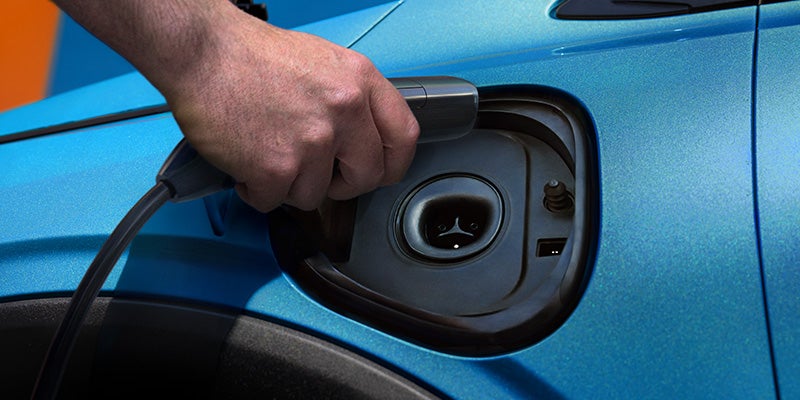 Close-up of a person's hand plugging a charging cable into the charging port of a bright blue electric vehicle.