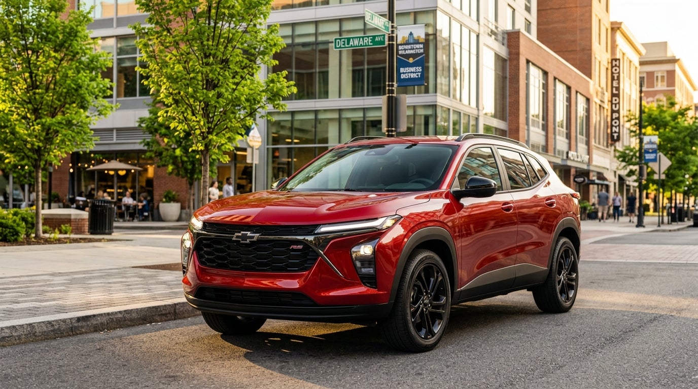 A photorealistic commercial automotive shot of a 2026 Chevrolet Trax in vibrant Apex Red metallic paint, parked on a clean sun-drenched street in a modern Northeastern Wilmington Delaware American downtown district, low-angle three-quarters front view, highlighting the sleek aerodynamic body lines a