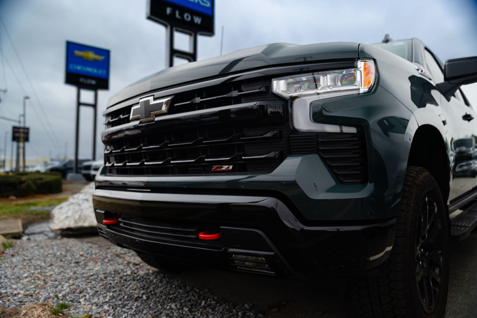 Close-up of the front grille and headlight of a dark green Chevrolet Silverado parked at a dealership, with dealership signs blurred in the background.
