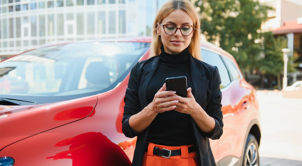Woman using her phone while standing in front a red vehicle.