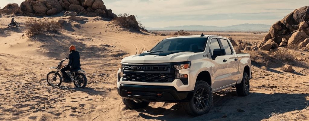 A white 2024 Chevy Silverado 1500 Custom parked beside a motorcycle rider.