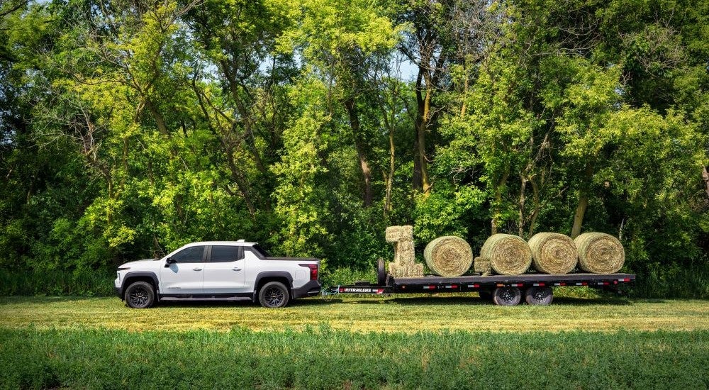 A white 2024 Chevy Silverado EV WT parked on a grass field with a trailer of hay bales.
