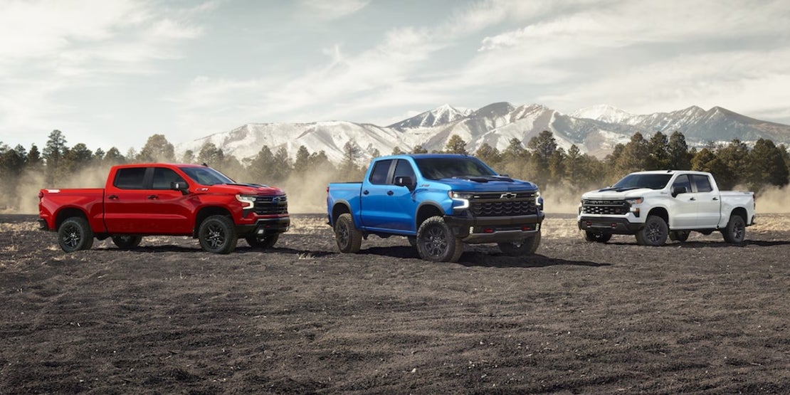 3 2026 Chevrolet Silverado pickup trucks, one red, one blue, and one white, parked on a dirt field with mountains in the background