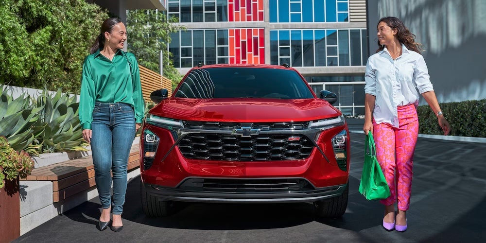 Tow women walking on either side of a red 2026 Chevrolet Trax SUV