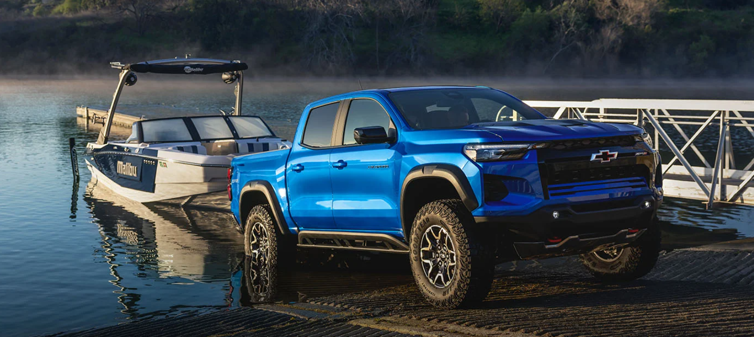 A blue pickup truck Colorado is parked on a boat ramp near a lake. It's towing a white boat labeled "Malibu" by a metal dock. The scene feels calm and adventurous.