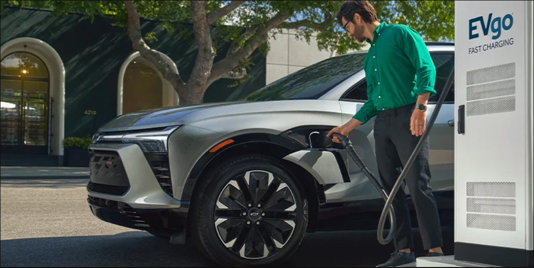 A man in a green shirt charges an electric vehicle at an EVgo fast charging station. The Chevy-Blazer-EV is silver, parked under a tree in a sunlit area.